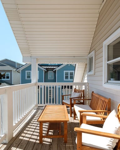 Covered deck with wooden chairs and table, overlooking blue and white residential buildings under clear sky