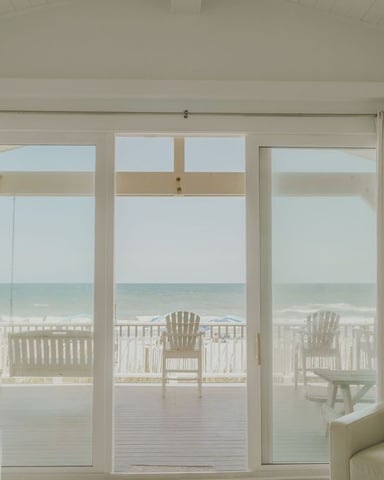 Bright living room with cream-colored furniture and large glass doors opening to a beachfront deck with ocean view