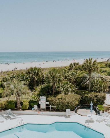 Swimming pool with lounge chairs overlooking beach, dunes, palm trees, and ocean