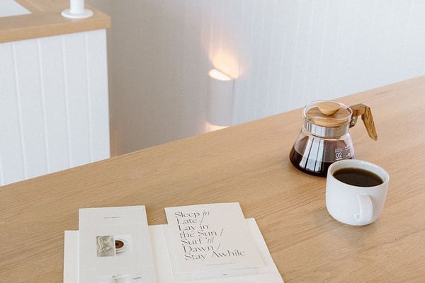 Wooden table with coffee cup, glass carafe, and open magazines in bright, minimalist room with white walls