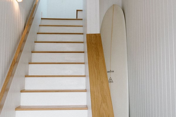 Staircase with white risers and wooden treads, wooden handrail, surfboard leaning against wall, and framed botanical art above.