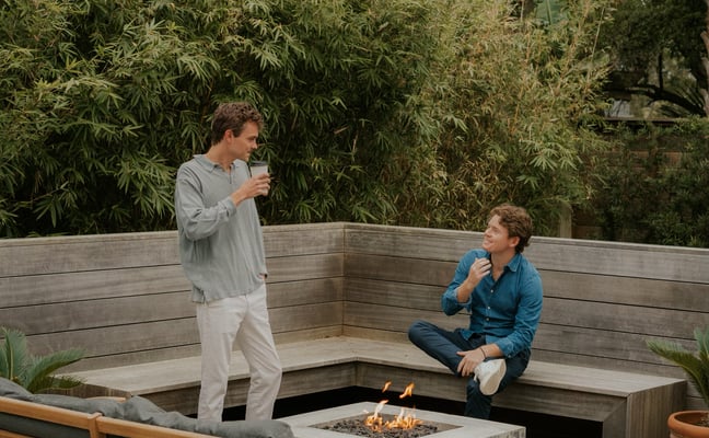 Two people on a wooden deck with built-in seating around a fire pit, surrounded by lush tropical plants and palm trees.